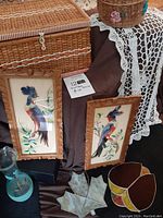 Two framed vintage Mexican feather bird artworks with colorful feathers in wooden carved frames, a light blue glass vase, and stained glass piece are visible on a cloth-covered surface along with a wicker basket and a crochet cloth.