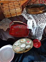Lot overview showing pink Pyrex bowl, red drip glaze ceramic bowl with lid, souvenir trays, woven basket, and pressed glass dish on table
