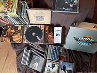 Photo showing the open trivia game box, some of the game components, and a pile of assorted music CDs in cases and sleeves.