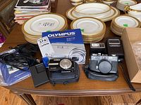 Gaf Memo 35EE camera front and Olympus Stylus 830 camera with manual and charger shown together on table with plates in background