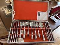 Opened case showing the complete set of knives, forks, spoons, and teaspoons arranged inside a fitted orange velvet case.
