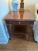Side view of one Deilcraft wooden side table showing drawer front with keyhole and dovetail joints, lower shelf, tapered legs with groove detail, placed next to wall and couch on hardwood floor.