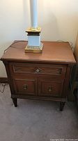 Front view of one wooden side table with a drawer on top and two cabinet doors below, medium brown finish, decorative metal knobs, topped with a lamp base without shade.