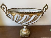 Front side angle of the oval white porcelain bowl featuring gold hand painted leaf swirls and decorative brass handles on each end with a detailed metal base.