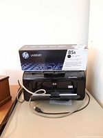 Black HP LaserJet P1102w printer with USB cable and HP 85A toner cartridge box on top, on beige table against wall.