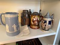 Shelf photo showing four items: light blue Wedgwood style pitcher, wooden stein with metal lid, ceramic steins with painted and relief designs