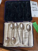 Set of six sterling silver spoons arranged inside a fitted padded case with black velvet and cream fabric lining.