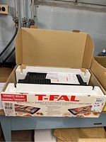 Original cardboard box with T-Fal branding visible, showing adjustable thermostat and indoor electric barbecue features in bilingual text.