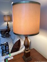 Pair of trilight table lamps with beige fabric shades on wooden side table beside sofa with brown pillows. Lamps are lit showing warm beige glow.