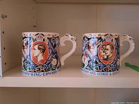 Two vintage ceramic Coronation mugs placed side by side on shelf showing front designs with royal profiles and crowns