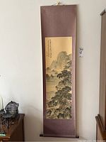 Full view of the vertical Asian wall scroll hung on wall showing the painted landscape and calligraphy mounted on a reddish-brown fabric backing with wooden rod at bottom.