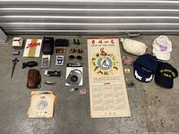 Photo showing a collection of hats, a 1998 zodiac calendar, sunglasses, boxes of playing cards, a vintage camera, small bottles, and assorted small items laid out on a concrete floor.