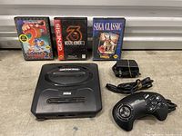 Sega Genesis console with controller, power adapter and three game cartridges: Sonic the Hedgehog 2, Mortal Kombat 3, The Revenge of Shinobi.