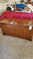 Front view of wooden cedar chest on carpet with folded blankets on nearby bed, showing paneled front with slight wear.