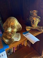 Two brass-finished metal clown bookends placed on a wooden shelf, one with a rounded clown nose and the other with a bow tie and hat.