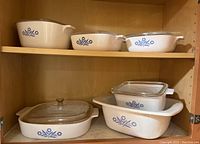 Seven pieces of Corning Ware Blue Cornflower bakeware arranged inside a wooden cabinet. Various sizes of ceramic dishes with some glass lids.