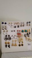 Full display of 24 pairs of mostly stud and dangle costume jewelry earrings arranged in rows on white backing board.