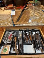 Photo of wooden knife block holding four knives and black drawer organizers with kitchen utensils and cutlery on granite countertop.