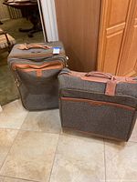 Two Hartmann luggage pieces made of grey tweed canvas with brown leather accents placed on floor.