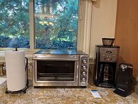 Front view of KitchenAid convection oven, Cuisinart coffeemaker, electric can opener and paper towel holder on granite countertop near window.