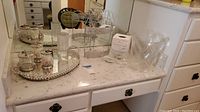 Full view of powder room accessories on counter including mirror tray, bathroom canisters with silver lids, tissue box cover, soap dish, clear acrylic trash can, and metal rack