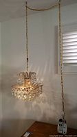 Photo showing swag chandelier hanging from ceiling via a gold chain, illuminating with a warm light, placed near a window and table.