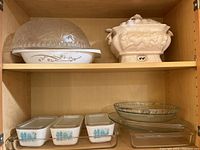Shelf showing beige ceramic tureen with ladle and a clear glass covered casserole dish on top shelf, three Pyrex blue rooster refrigerator dishes with lids on bottom shelf along with additional clear glass dishes.