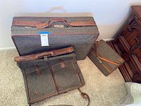 Three Hartmann luggage pieces arranged on carpet: a large suitcase, a smaller box case, and a garment bag lying flat showing front pockets