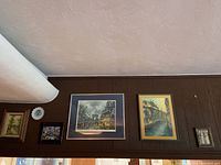 Wide view of wall with several framed art pieces and decorative plate hung high on tan wood paneling wall.