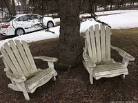 Pair of white painted wooden Adirondack chairs outside near a tree, showing weathered and worn paint.