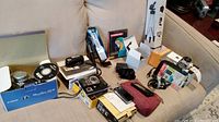 Digital cameras and accessories spread on couch including boxed cameras, GPS device in case, tripod box, memory card adapter boxes.