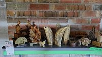 Photo shows a variety of carvings made of different materials including wood, bone or ivory-like objects, and possibly stone pieces arranged on a surface against a brick wall.