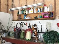 Wide view of gardening supplies on shelf and workbench including watering cans, hand tools, bottles of plant food and bug spray, and artificial foliage.