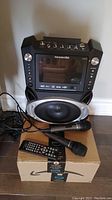 Front view of Karaoke USA machine showing built-in screen, speaker, two microphones, remote, and cables on a box.