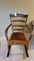 Front view of vintage wood and wicker rocking chair showing curved wooden arms, woven wicker back, and brown seat cushion.