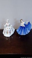 Two Royal Doulton figurines side by side, Elaine in white floral gown and Diana in blue dress