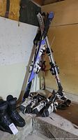 Ski equipment including skis, poles, and several boots and skates grouped together in a basement corner.