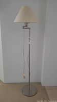 Full view of the metal floor lamp showing round base, vertical pole, extension arm, hanging pull cord, and beige lampshade.