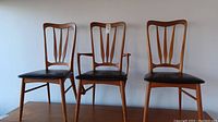 Three teak dining chairs in a row against a white wall: one armchair centered between two side chairs
