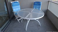 Full view of round glass top patio table with two blue cushioned chairs on balcony.