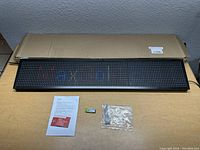 Front view of LED programmable scrolling sign laid on a cardboard surface, accompanied by USB drive, instruction paper, and a plastic-adapted cable.