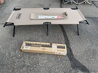 Photo of the tan Coleman camping cot on black metal frame with carry bag and cardboard box on pavement ground.