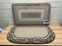 Photo showing both floor mats; rectangular mat with geometric pattern positioned vertically on wall, rounded mat with border pattern laid on table.