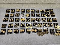 Photo showing 50 pairs of assorted gold-tone earrings on black branded card backing, laid out on a gray surface against a white wall.