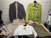 Five jackets laid out on a wooden surface showing various colors and styles including gray, lime green, beige, white, and olive green.