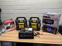 Lot of 5 battery chargers and jump starters arranged on wood table against cinder block wall. Visible items include 2 DEWALT jump starters, VECTOR charger, small boxed Schumacher charger, and small black charger unit.