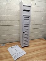Full view of white metal bathroom tissue holder cabinet and instruction sheet on wooden table in front of cinder block wall.