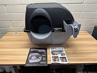 Front view of Omega Paw Roll & Clean self-cleaning cat litter box with waste bin on the side, placed on wooden table with two instruction manuals.