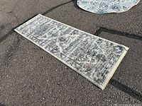 Top side view of rectangular runner rug with a gray and white patterned design lying on asphalt outdoor surface.