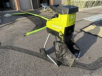 Side-angle photo of the green and black Sun Joe wood chipper showing the feeding chute, frame, wheels, and attached black fabric grass catcher bag.